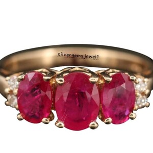 Puede incluir: Un anillo de oro con tres rubíes rojos vibrantes de corte ovalado en fila, flanqueados por pequeñas piedras transparentes a cada lado. La banda es de oro liso y pulido. El texto silvergemsjewel1 está impreso en la superficie interior.