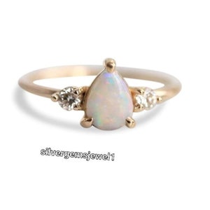 Puede incluir: Un anillo de oro con una piedra central de ópalo en forma de pera, flanqueada por dos piedras preciosas redondas y transparentes más pequeñas. El anillo tiene una banda delicada y un diseño sencillo. El texto "silvergemsjewel1" es visible en la parte inferior.