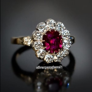 Puede incluir: Un anillo de estilo vintage con un gran rubí de forma ovalada rodeado por un halo de diamantes más pequeños. El anillo está engastado en plata y tiene una banda delicada.