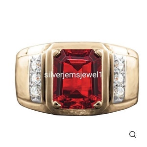 Puede incluir: Un anillo dorado con una gran gema roja rectangular en el centro. El anillo está adornado con pequeñas gemas redondas y claras a cada lado de la piedra central. El texto "silverjemsjewel1" es visible en el anillo.