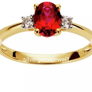 Puede incluir: Un anillo de oro con una piedra roja ovalada en el centro, flanqueada por dos piedras redondas y transparentes más pequeñas. La banda del anillo es dorada y las piedras están engastadas en oro. Joya.