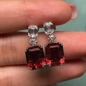 Puede incluir: Un par de pendientes colgantes con gotas de piedras preciosas rojas y detalles de diamantes. Los pendientes presentan una piedra preciosa roja de forma cuadrada con un diamante encima y un diamante más pequeño debajo.