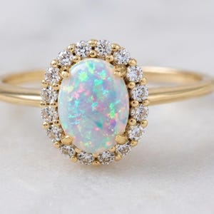 Puede incluir: Anillo de oro con una piedra de ópalo ovalada rodeada de pequeños diamantes. El ópalo muestra colores iridiscentes. La banda del anillo es de oro pulido. Una joya.