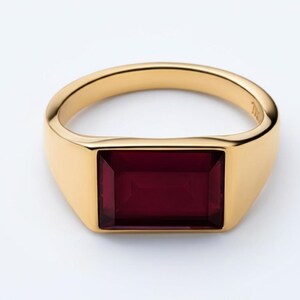 Puede incluir: Un anillo dorado con una gema rectangular de color rojo intenso. El anillo tiene un acabado liso y pulido y un estilo clásico de sello. La gema está empotrada en el diseño del anillo.