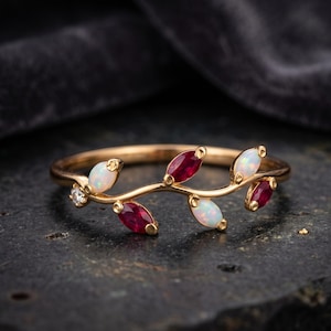 May include: A gold ring featuring a delicate vine-like band adorned with marquise-cut red rubies and oval-shaped white opals. A single small round clear gemstone is set at the base of the vine design, creating a symmetrical botanical arrangement.