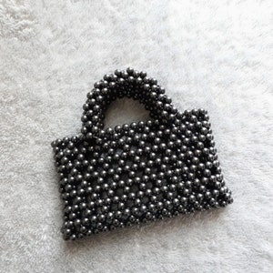 Pode incluir: Uma bolsa de mão preta de contas com alça superior. A bolsa é feita de pequenas contas redondas que são tecidas juntas para criar uma superfície texturizada.