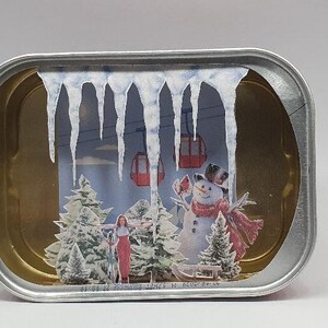 Puede incluir: Una lata de metal plateada con una escena de invierno en su interior. La escena incluye un muñeco de nieve, una pareja de personas y un telesilla. La escena está enmarcada por carámbanos que cuelgan de la parte superior de la lata.