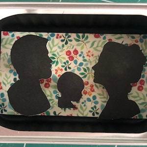 Puede incluir: Un retrato familiar en silueta, con un padre, una madre y un niño, recortados en papel negro y colocados sobre un fondo con estampado floral.