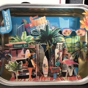 Puede incluir: Un diorama en miniatura dentro de una lata que representa una escena de playa con palmeras, un flamenco rosa, una tabla de surf, un coche y un cartel que dice "Ocean Dr".