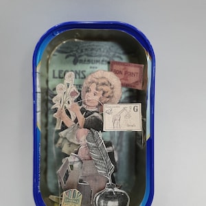 Puede incluir: Una obra de arte de técnica mixta en una lata azul. La obra presenta un collage de imágenes vintage, incluyendo una niña, flores, una pluma y un tintero. Las palabras "BON POINT" y "GIRAFE" son visibles.