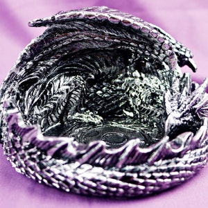 May include: A silver dragon figurine with its wings spread out, forming a bowl shape. The dragon is detailed with scales and has a textured surface.