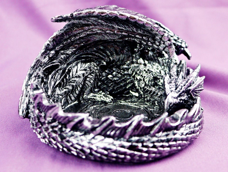 May include: A silver dragon figurine with its wings spread out, forming a bowl shape. The dragon is detailed with scales and has a textured surface.