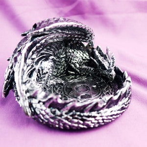 May include: A silver dragon figurine shaped like a bowl. The dragon is curled up with its head resting on its tail, creating a circular shape. The dragon's scales are detailed and the figurine has a shiny finish.