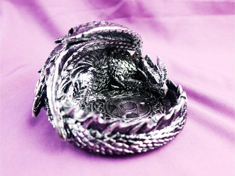 May include: A silver dragon figurine shaped like a bowl. The dragon is curled up with its head resting on its tail, creating a circular shape. The dragon's scales are detailed and the figurine has a shiny finish.