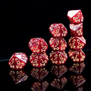 May include: A stack of red and gold polyhedral dice, arranged in a stepped pyramid. The dice feature gold numbering and detailed designs. The image is set against a black background, with reflections of the dice visible.