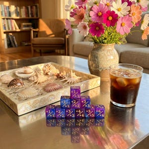 May include: A set of purple glittery dice arranged in a pyramid, each marked with numerical values and plus signs. The dice are positioned on a metallic surface next to a square tray filled with various seashells and a glass of iced coffee.