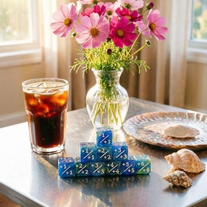 May include: A set of blue and green marbled dice arranged in a pyramid, each marked with numerical fractions. Beside the dice are a glass of dark iced coffee, a clear glass vase filled with pink cosmos flowers, and two seashells on a plate.