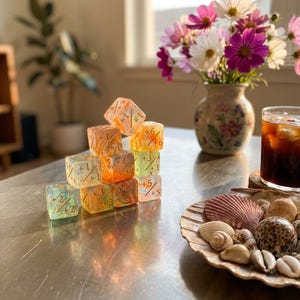 May include: A stack of translucent, multi-coloured polyhedral dice featuring gold-coloured numbers and mathematical symbols. The dice display various hues including orange, yellow, and light green, arranged in a pyramid formation on a smooth surface.