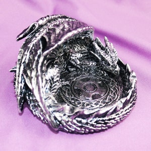 May include: A silver dragon figurine with intricate details, curled into a ring shape, with a circular design in the center. The dragon's wings are spread out and its head is facing the viewer.