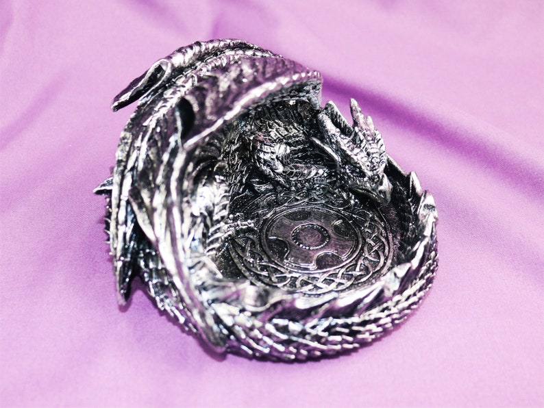 May include: A silver dragon figurine with intricate details, curled into a ring shape, with a circular design in the center. The dragon's wings are spread out and its head is facing the viewer.