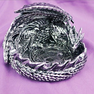 May include: A silver dragon figurine with its wings spread, curled into a circular shape, forming a decorative dish. The dragon's head is visible, and the dish has a circular design in the center.