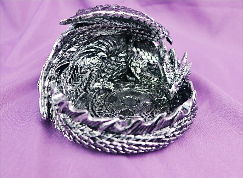 May include: A silver dragon figurine with its wings spread, curled into a circular shape, forming a decorative dish. The dragon's head is visible, and the dish has a circular design in the center.