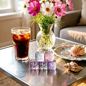 May include: A glass of iced coffee, a clear glass vase filled with pink and white cosmos flowers, two seashells, and five translucent purple and blue dice with white fraction markings ranging from 1/1 to 5/5 arranged on a metal table.