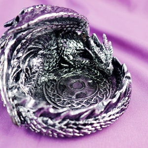 May include: A silver dragon figurine with a detailed design, curled into a circular shape to form a dish. The dragon's wings and tail are intricately sculpted, and the dish features a Celtic knot design in the center.