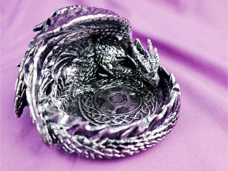 May include: A silver dragon figurine with a detailed design, curled into a circular shape to form a dish. The dragon's wings and tail are intricately sculpted, and the dish features a Celtic knot design in the center.