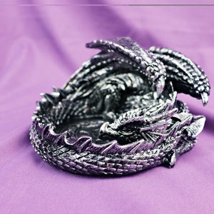 May include: A silver dragon figurine curled up in a sleeping position. The dragon has detailed scales and wings. The figurine is perfect for a fantasy-themed home decor.