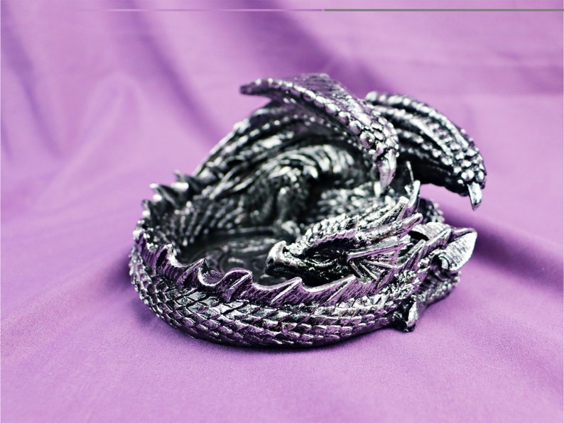 May include: A silver dragon figurine curled up in a sleeping position. The dragon has detailed scales and wings. The figurine is perfect for a fantasy-themed home decor.