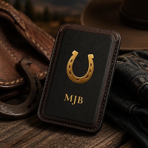 May include: A black and brown leather phone case with a gold horseshoe emblem and the initials "MJB". The case is displayed on a wooden surface with a cowboy hat, denim jacket, and a leather bag in the background.