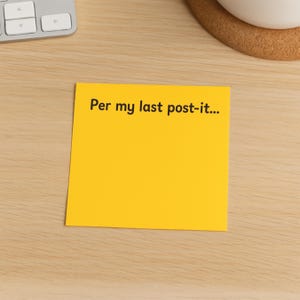 May include: A bright yellow Post-it note with the text "Per my last post-it..." in black. The sticky note is on a light wood surface, with a keyboard and a white mug visible in the background.