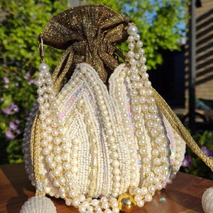 May include: An off-white beaded pouch bag with a gold drawstring closure and pearl accents. The bag features a gold strap and a tassel. The bag is sitting on a wooden surface.