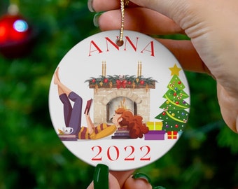 Puede incluir: Un adorno navideño de cerámica blanca con el nombre "ANNA" en letras rojas. El adorno presenta una ilustración festiva de una chimenea, un árbol de Navidad y regalos, con el año "2022" en rojo. Una cuerda dorada está unida para colgar.