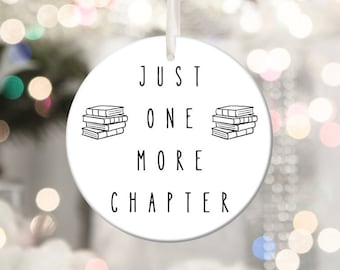 May include: White ceramic Christmas ornament with black text that reads "Just One More Chapter" and a black and white illustration of a stack of books.