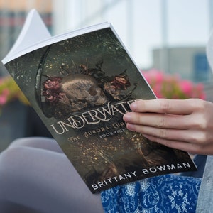 May include: A person is reading a paperback book titled "Underwater: The Aurora Chronicles, Book One" by Brittany Bowman. The cover features a dark, gothic illustration of a skull with flowers and vines.