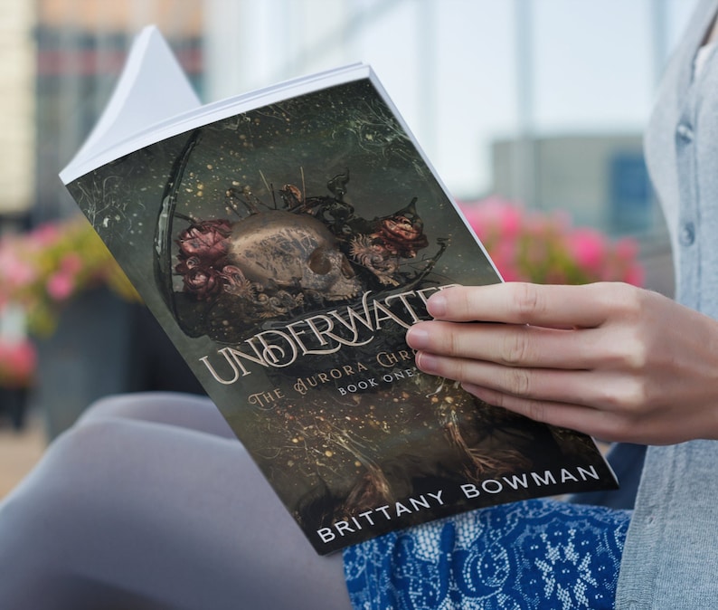 May include: A person is reading a paperback book titled "Underwater: The Aurora Chronicles, Book One" by Brittany Bowman. The cover features a dark, gothic illustration of a skull with flowers and vines.