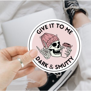May include: A pink and white round sticker featuring a skull wearing a pink beanie and holding a coffee cup. The text on the sticker reads "Give it to me dark & smutty".
