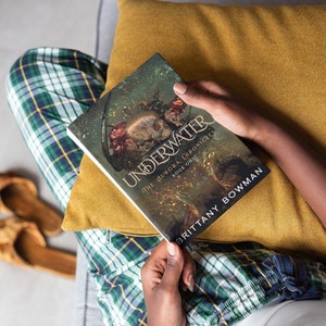 May include: A person is reading a book titled "Underwater: The Aurora Chronicles, Book One" by Brittany Bowman. The book cover features a dark, fantasy-inspired illustration with a skull and flowers.