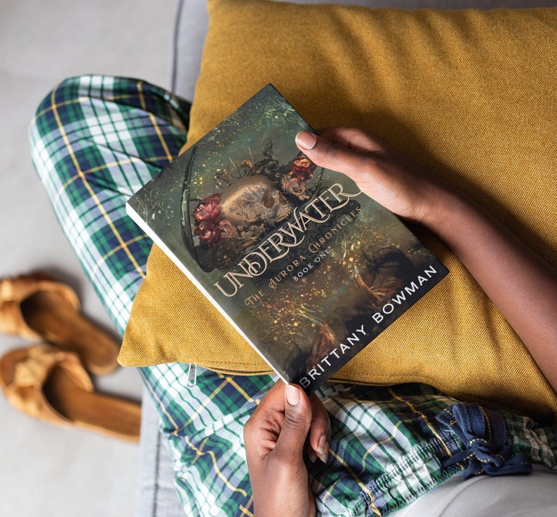 May include: A person is reading a book titled "Underwater: The Aurora Chronicles, Book One" by Brittany Bowman. The book cover features a dark, fantasy-inspired illustration with a skull and flowers.
