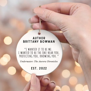 May include: Round white ceramic ornament with gold hanging string. The ornament displays text: "AUTHOR BRITTANY BOWMAN" and a quote from "Underwater: The Aurora Chronicles." It is dated EST. 2022.