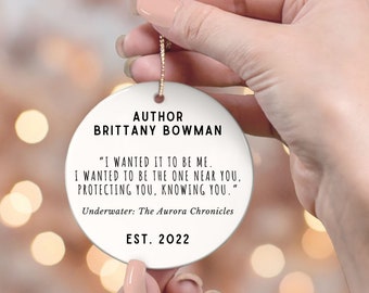 May include: White ceramic ornament with gold string, featuring text: "AUTHOR BRITTANY BOWMAN" and a quote from "Underwater: The Aurora Chronicles." The ornament is dated EST. 2022.