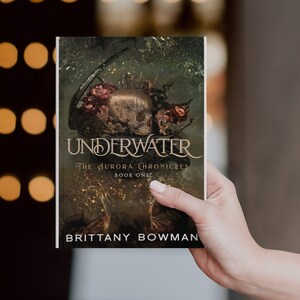 May include: A book cover with a dark green background and a skull with flowers on top. The title is "UNDERWATER" and the subtitle is "THE AURORA CHRONICLES BOOK ONE". The author's name is "BRITTANY BOWMAN".