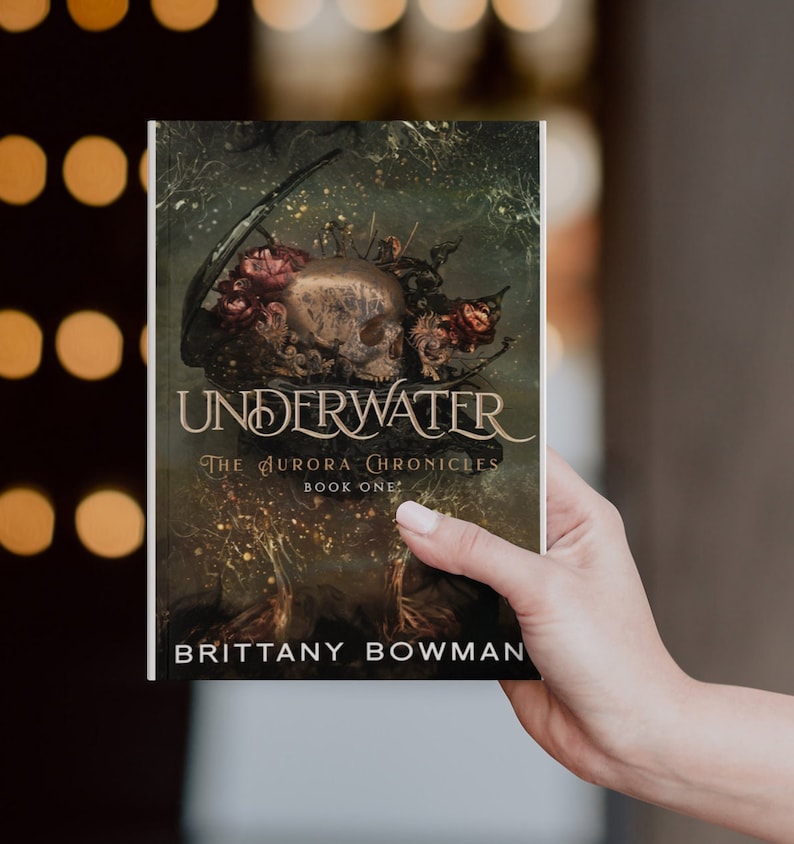 May include: A book cover with a dark green background and a skull with flowers on top. The title is "UNDERWATER" and the subtitle is "THE AURORA CHRONICLES BOOK ONE". The author's name is "BRITTANY BOWMAN".