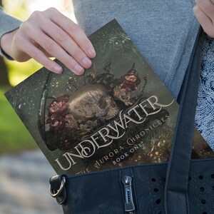 May include: A paperback book with a dark green cover featuring a skull with intricate details and a floral design. The title of the book is "Underwater" and the subtitle is "The Aurora Chronicles Book One".