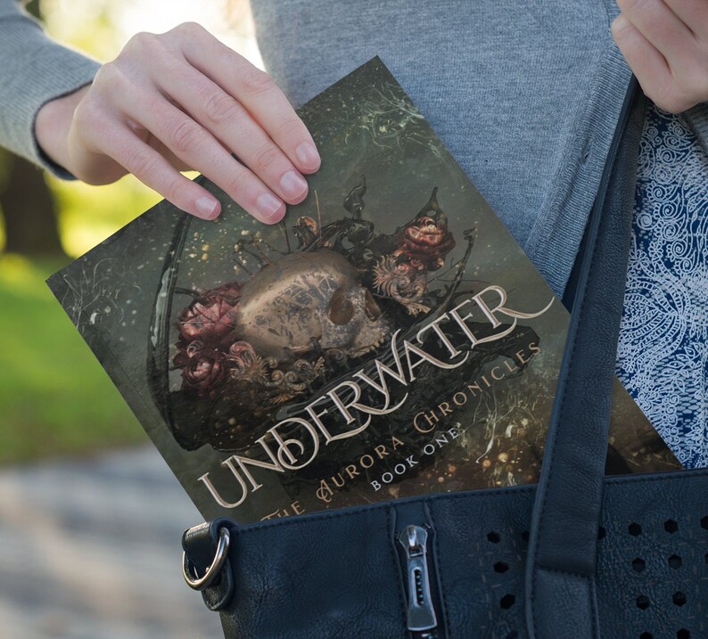 May include: A paperback book with a dark green cover featuring a skull with intricate details and a floral design. The title of the book is "Underwater" and the subtitle is "The Aurora Chronicles Book One".