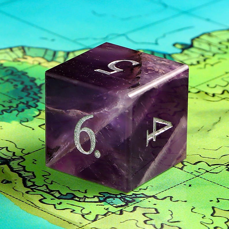 May include: A purple six-sided dice with silver numbers 1, 4, 5, and 6. The dice is made of amethyst and has a natural, rough texture.