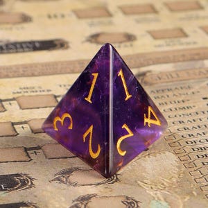 May include: A purple four-sided dice with gold numbers 1, 2, 3, and 4. The dice is made of amethyst and has a natural, faceted appearance.