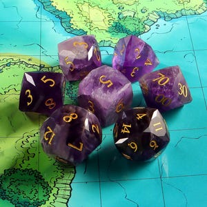 Può includere: Un set di sette dadi di pietre preziose viola con numeri dorati, disposti su una mappa. I dadi sono di varie forme poliedriche, tra cui un d4, d6, d8, d10, d12 e d20. I dadi sono di colore viola scuro.